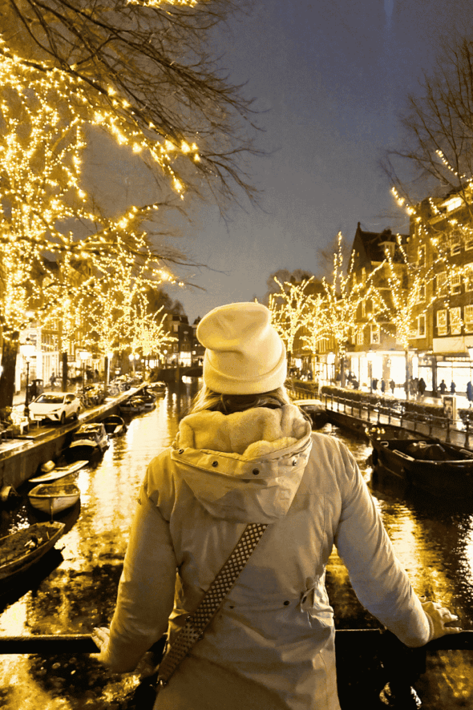 Kate stands on a bridge in Amsterdam at night, gazing down a canal lined with boats and trees glowing with golden holiday lights.