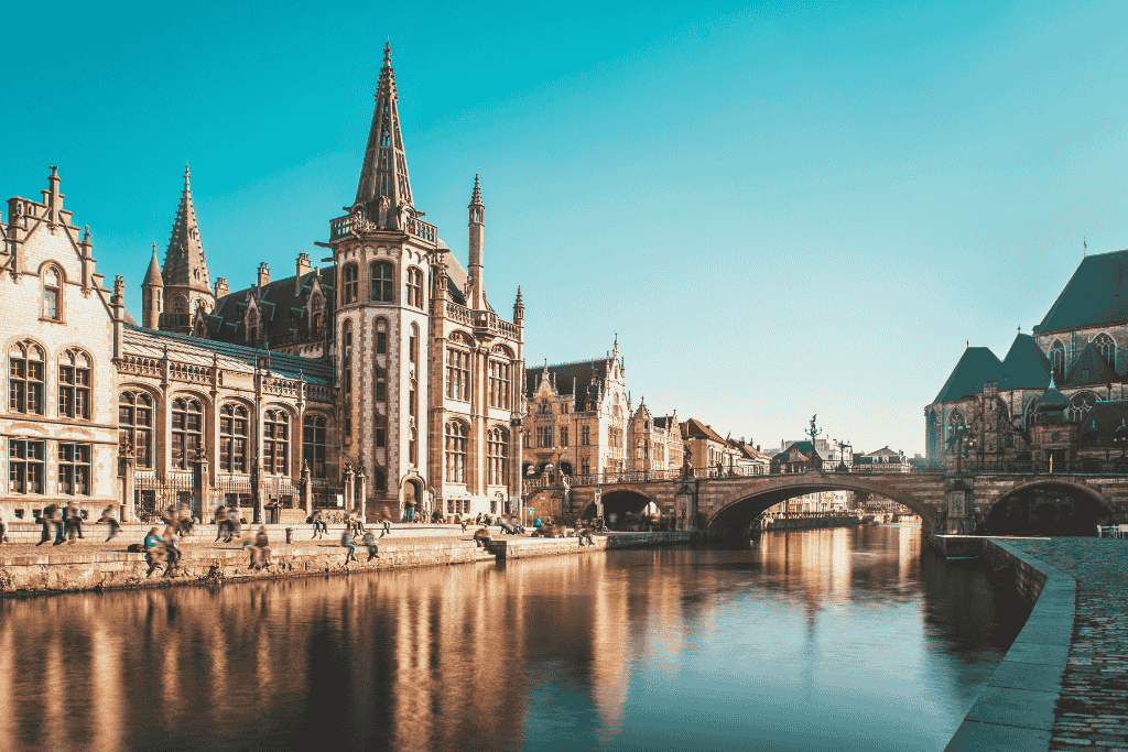 A serene canal in Ghent reflects the surrounding medieval buildings and arched stone bridge under a bright blue sky, with people strolling along the waterfront.