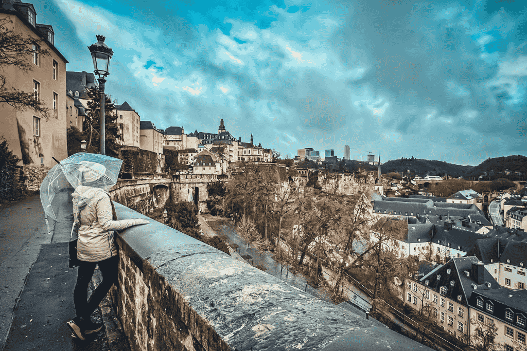 Kate, holding a transparent umbrella, looks out over the dramatic cliffs, historic buildings, and valley of Luxembourg City on a cloudy day.