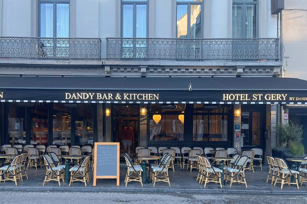 Street view of Boutique Hotel Saint-Géry in Brussels, with its cozy Dandy Bar & Kitchen terrace lined with wicker chairs and café tables under a black awning.