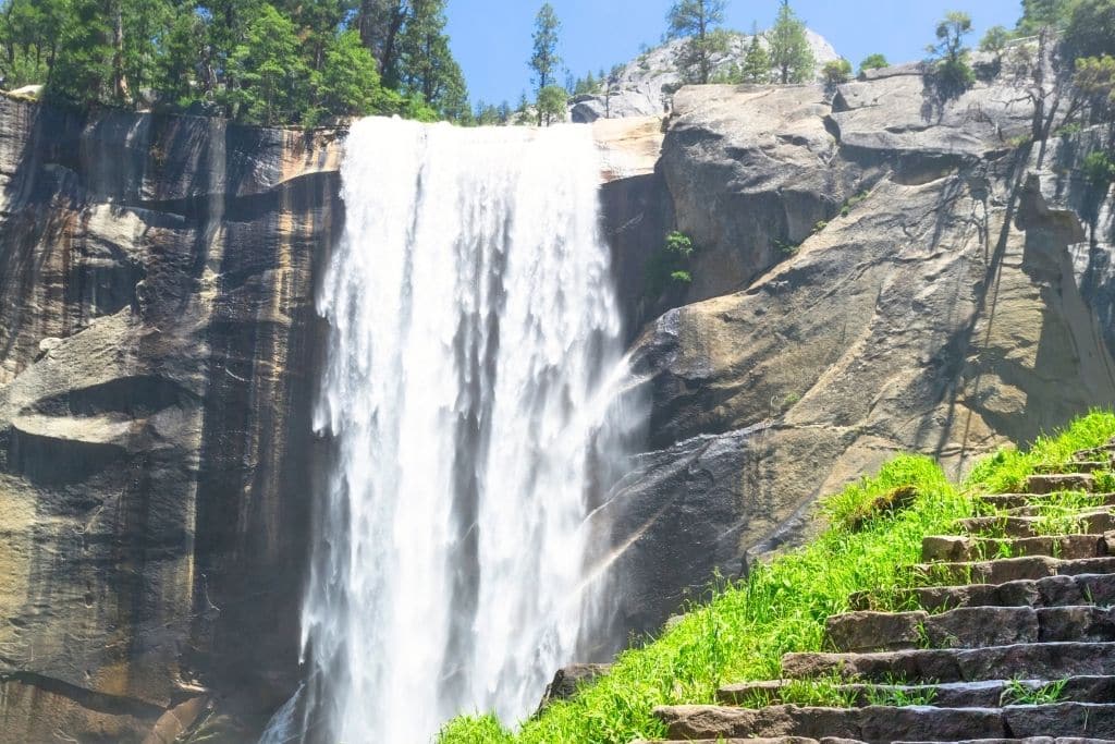 Vernal Fall plunges dramatically over a granite cliff, with bright green moss and stone steps in the foreground along the Half Dome Trail.