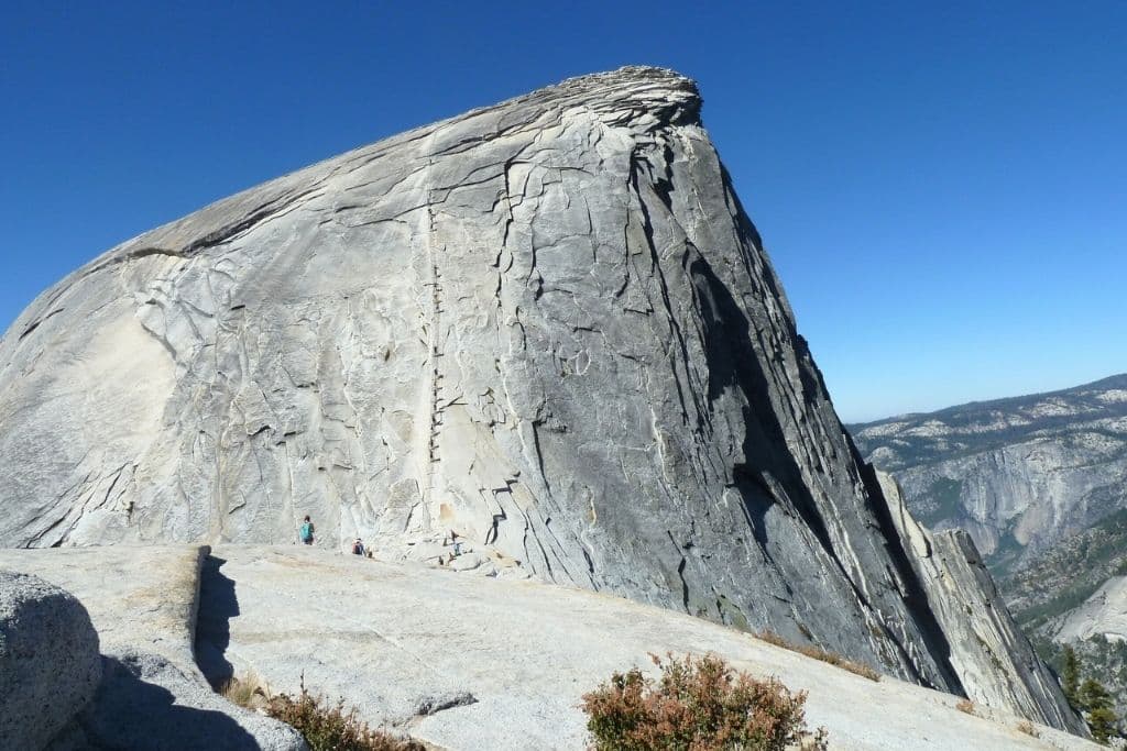 Hikers approach the base of Half Dome’s steep cable route, with the towering granite summit rising sharply under a clear blue sky.