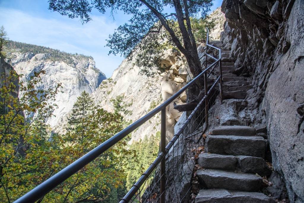 Narrow stone steps with a metal railing hug the cliffside along the Half Dome Trail, overlooking a forested canyon and distant granite cliffs.