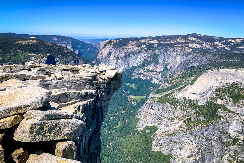 Sweeping view from the summit of Half Dome, showing its sheer granite edge overlooking Yosemite Valley and forested mountain ranges under a bright blue sky.