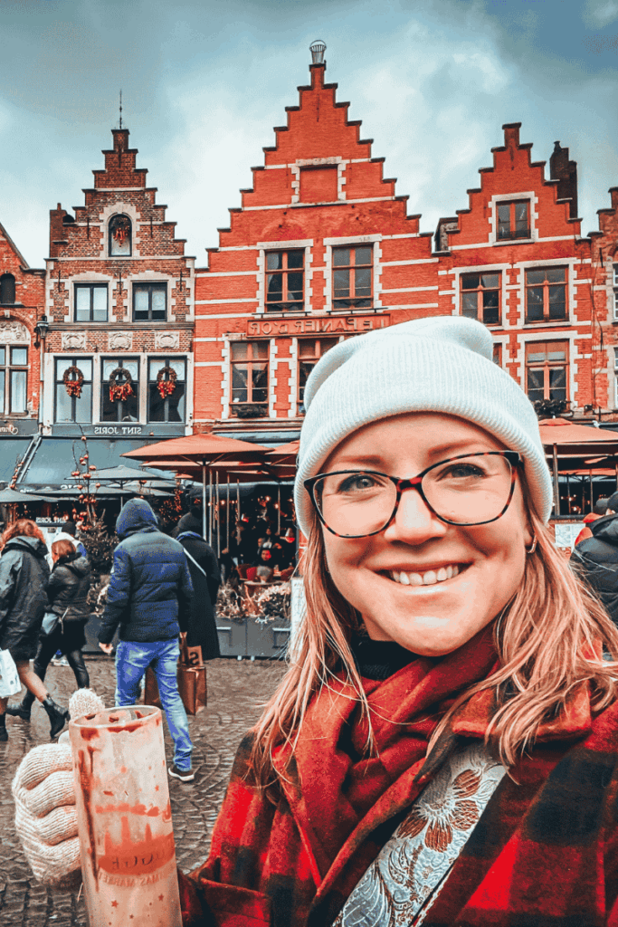 During a festive day trip from Brussels, Kate holds a tall mug of hot chocolate while smiling in Bruges’ town square, surrounded by red-brick gabled buildings.