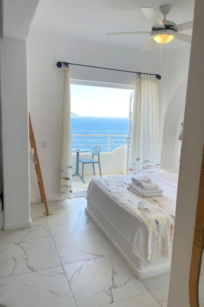 Bright, minimalist hotel room at Payam Hotel in Kaş, Turkey, featuring a bed with coastal-themed linens and a balcony overlooking the deep blue sea.