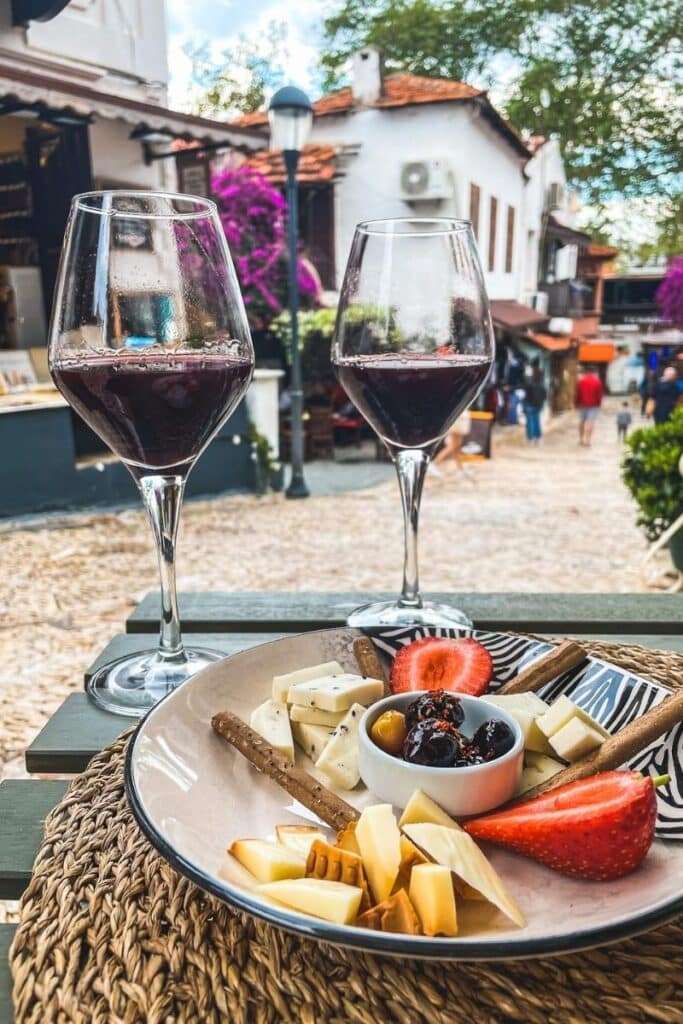 Two glasses of red wine sit beside a plate of Turkish cheeses, olives, strawberries, and breadsticks, set on an outdoor table in a charming Kaş street scene.