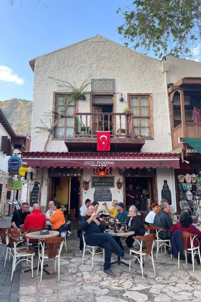 Locals and travelers enjoy drinks and conversation outside Old House Café & Bar in Kaş, Turkey, set in a rustic white-stone building with a Turkish flag and wood-framed windows.