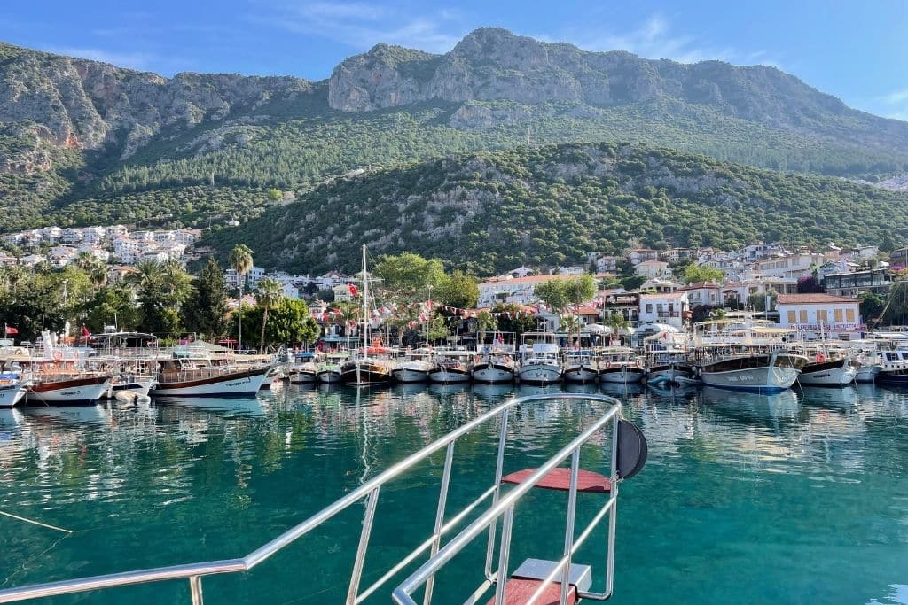Row of boats docked in the crystal-clear harbor of Kaş, Turkey, with whitewashed buildings and forested mountains rising in the background.