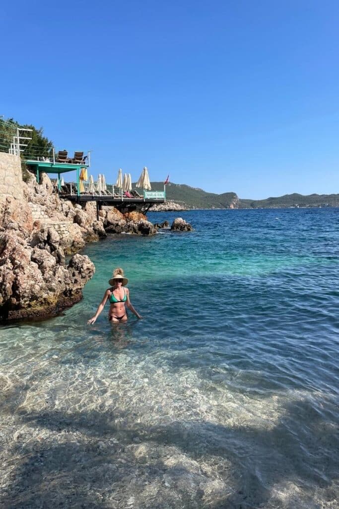 Kate wades in the clear blue waters near Derya Beach in Kaş, Turkey, with rocky shores and a sunbed-lined deck in the background.