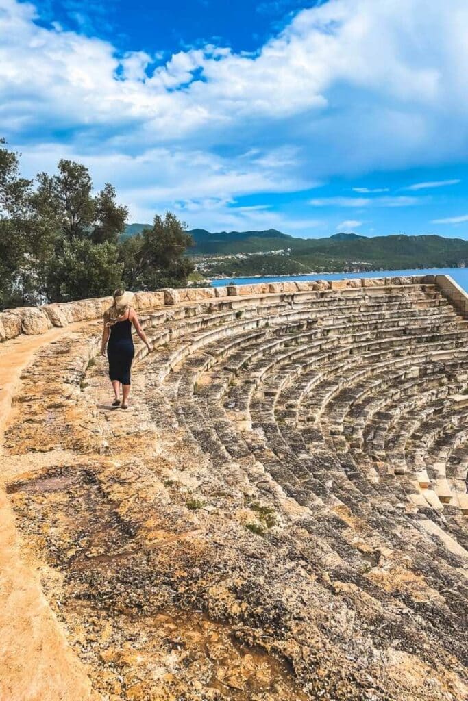 Kate in a sunhat and black dress walking along the top edge of the ancient amphitheater in Kaş, Turkey. The stone rows curve dramatically below her with a backdrop of green hills and blue sea under a partly
