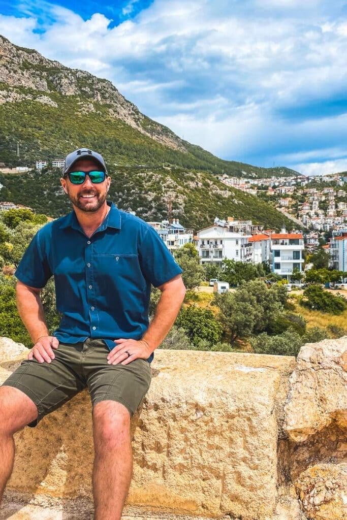 Kate's husband, wearing a navy shirt, green shorts, and sunglasses, sits on a stone wall with a view of Kaş, Turkey, in the background. Behind him are white buildings nestled into the green hillside beneath a partly cloudy sky.