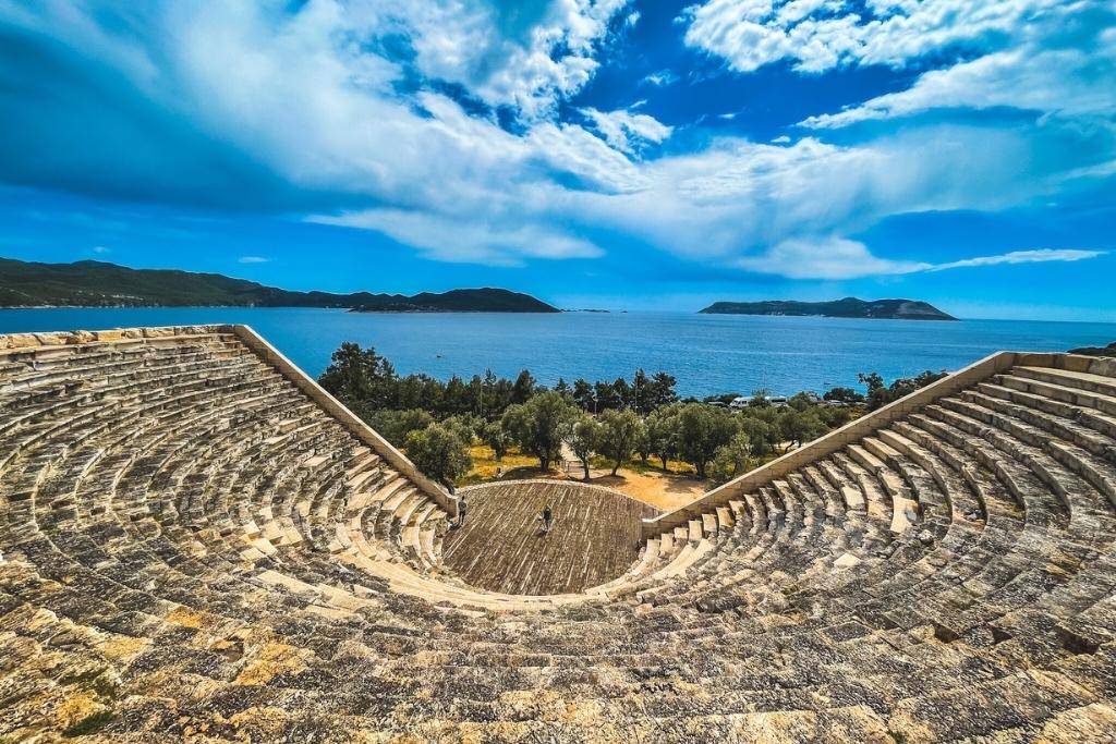 View from the top of the ancient amphitheater in Kaş, Turkey, with semi-circular stone seating overlooking a lush grove of trees and the brilliant blue Mediterranean Sea. Islands dot the horizon under a dynamic sky filled with sweeping clouds.