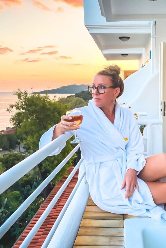 Kate relaxes on a balcony in a white robe, holding a glass of wine while watching the sunset over the Aegean coastline in Kaş, Turkey.