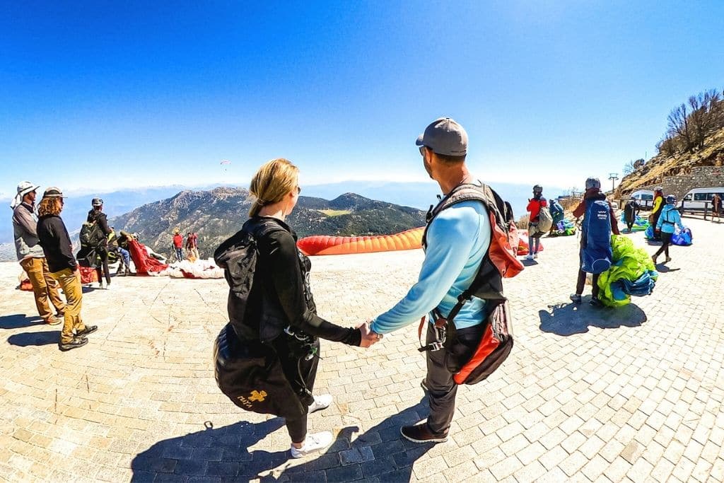 Kate and her husband hold hands at the launch site for paragliding in Ölüdeniz, Turkey, surrounded by fellow flyers and sweeping mountain views.