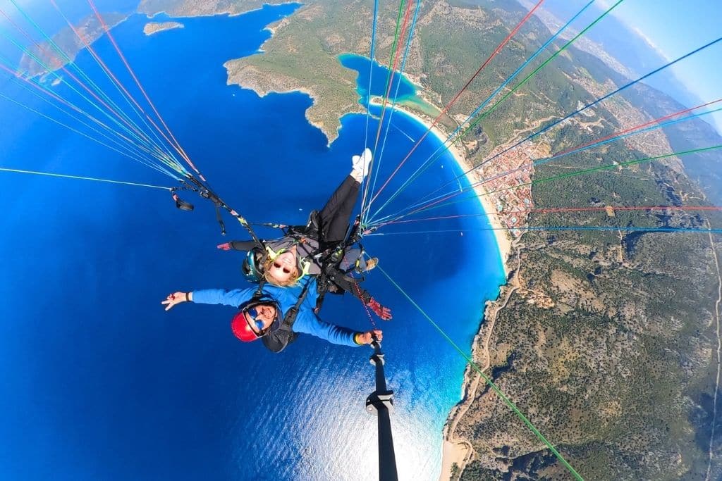 Kate paraglides high above the turquoise coast of Ölüdeniz, Turkey, with arms outstretched and panoramic views of the lagoon and surrounding mountains below.