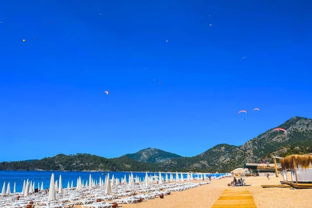 Rows of white sun loungers line the golden beach of Ölüdeniz, Turkey, as colorful paragliders soar above the mountains and sea under a bright blue sky.