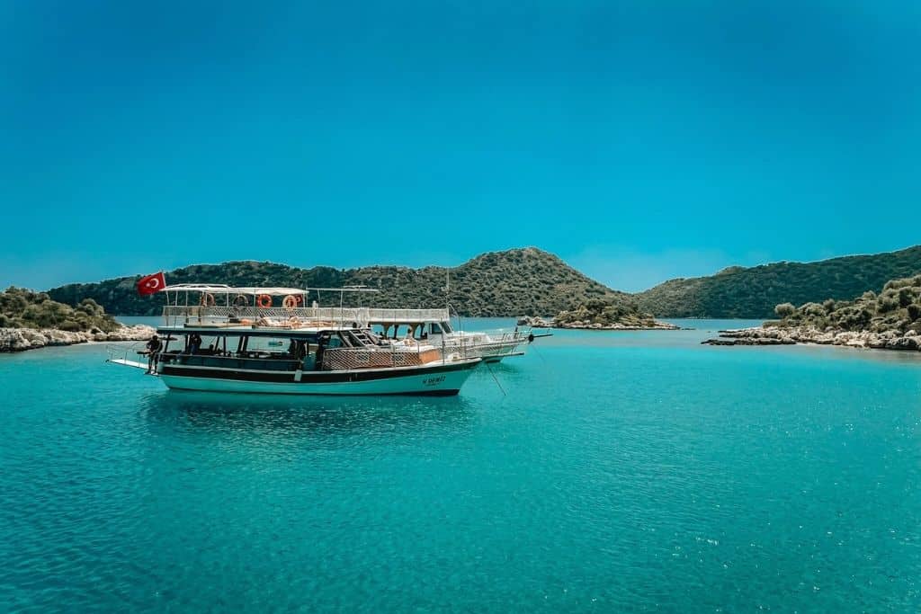 A traditional wooden boat with a Turkish flag floats on the vivid turquoise waters of the Turkish Riviera, surrounded by lush green islets under a clear blue sky.