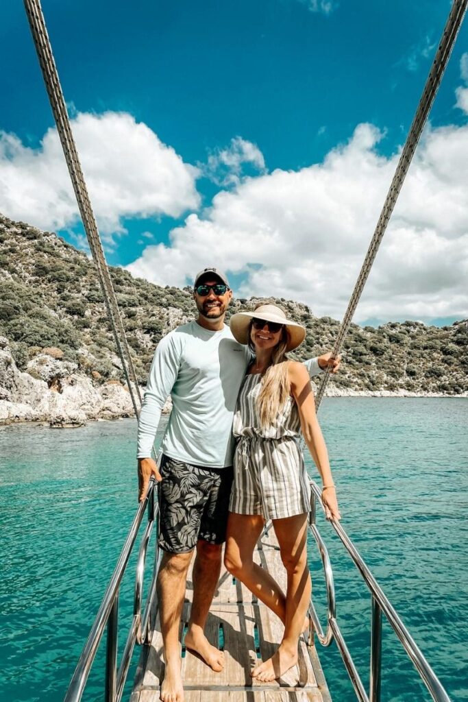 Kate and her husband smile while standing barefoot on the deck of a boat in the Turkish Riviera, with turquoise waters and lush rocky hills behind them.
