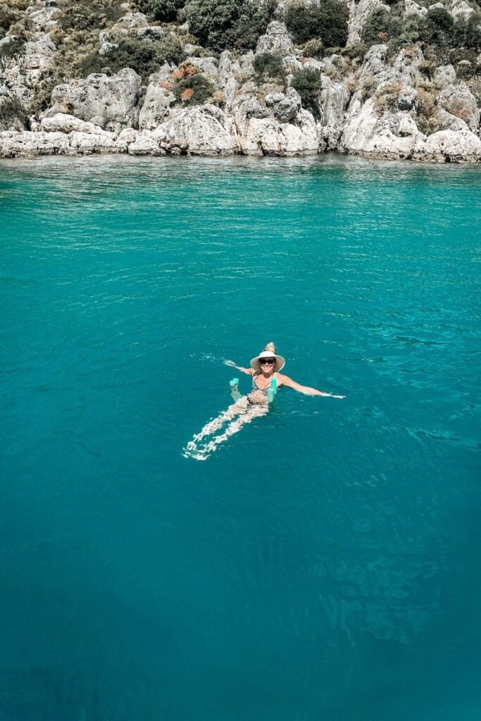 Kate floats in the clear turquoise waters of the Turkish Riviera, smiling beneath a sunhat with a rocky, green-covered shoreline in the background.