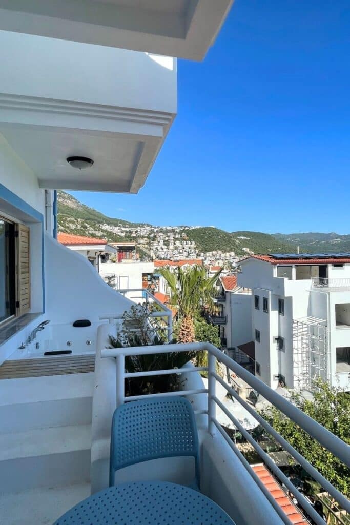 View from a hotel balcony in Kaş, Turkey, featuring a private jacuzzi, patio seating, and hillside homes nestled under a clear blue sky.
