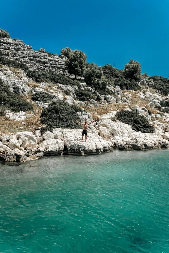 Kate’s husband stands with arms outstretched on a rocky ledge above turquoise water along the rugged coastline of the Turkish Riviera.