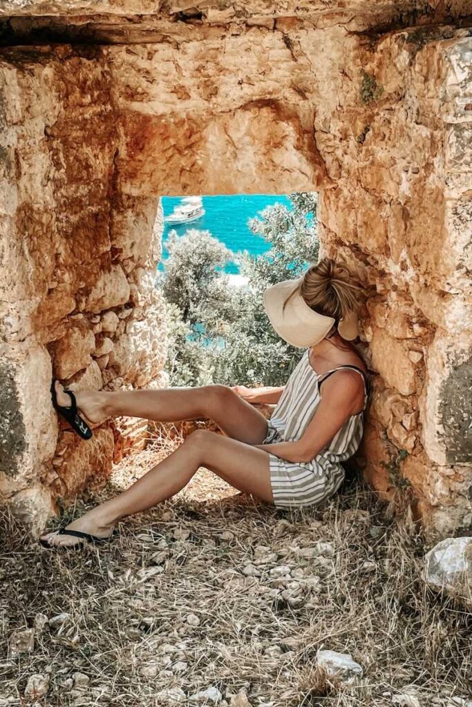 Kate lounges in a stone archway overlooking turquoise water and anchored boats along the Turkish Riviera, framed by ancient ruins and summer sun.