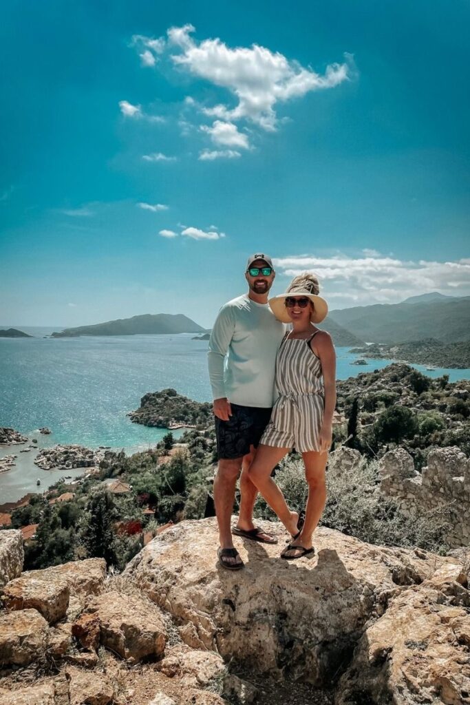 Kate and her husband pose on a rocky outcrop with panoramic views of the Mediterranean Sea and green hills near Kaş, Turkey.