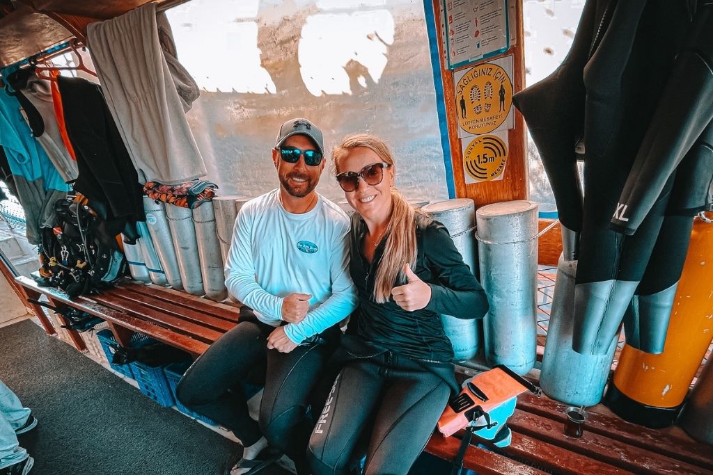 Kate and her husband sit on a dive boat in Kaş, Turkey, giving thumbs up in wetsuits beside scuba tanks and hanging gear, ready for a dive adventure.