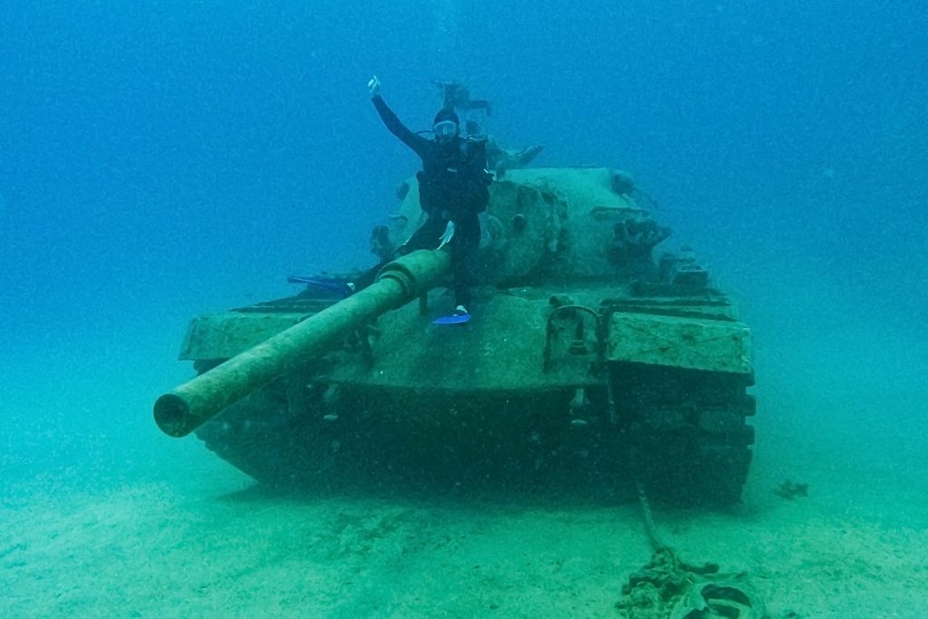 A scuba diver sits on the barrel of a sunken military tank at the bottom of the sea near Kaş, Turkey, giving a thumbs-up in the eerie blue depths.