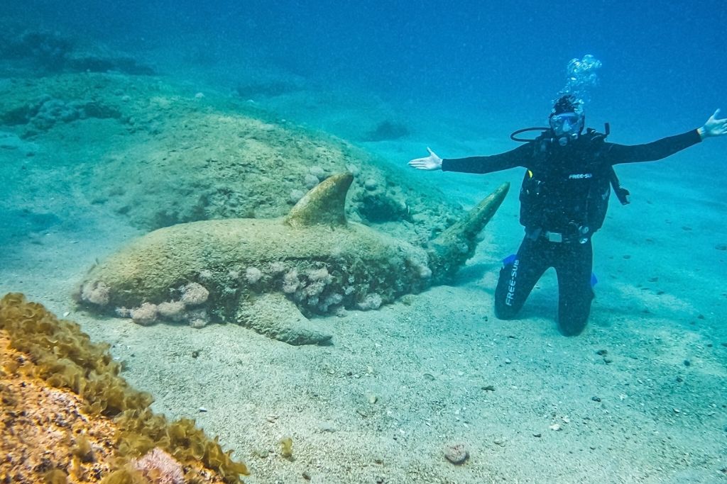 A scuba diver poses underwater beside a large stone shark sculpture encrusted with coral off the coast of Kaş, Turkey.