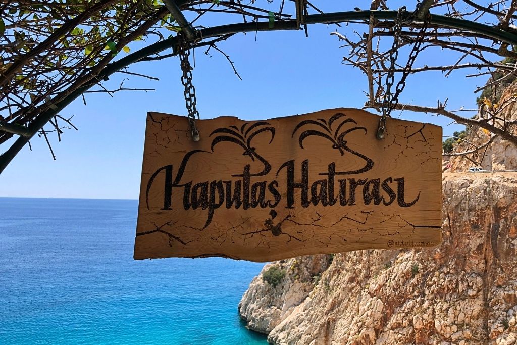 A wooden sign reading “Kaputaş Hatırası” (Kaputaş Memory) hangs from tree branches above turquoise Mediterranean waters and dramatic cliffside views near Kaş, Turkey.