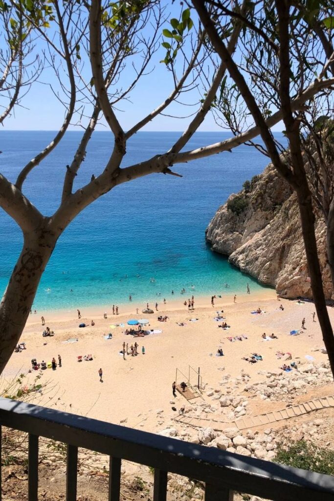 A view of Kaputaş Beach in Turkey framed by tree branches, showing golden sand, swimmers in turquoise water, and sunbathers spread along the shoreline.