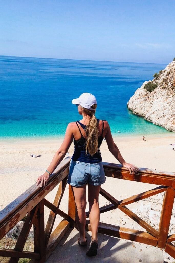 Kate gazes out over the vivid blue sea and golden sand of Kaputaş Beach from a wooden lookout point perched above the cliffs.