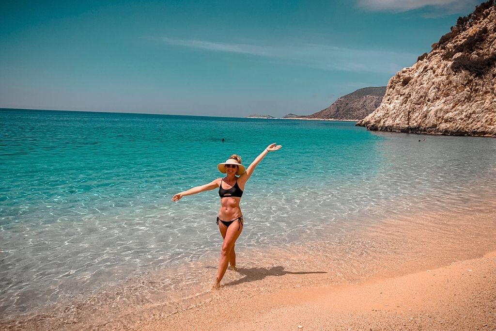 Kate strikes a joyful pose on the golden sand of Kaputaş Beach, with clear turquoise waves lapping at her feet and rugged cliffs in the background.