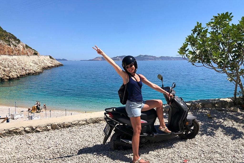 Kate poses on a scooter flashing a peace sign beside the sparkling turquoise coast near Kaputaş Beach, with rocky cliffs and clear skies in the background.