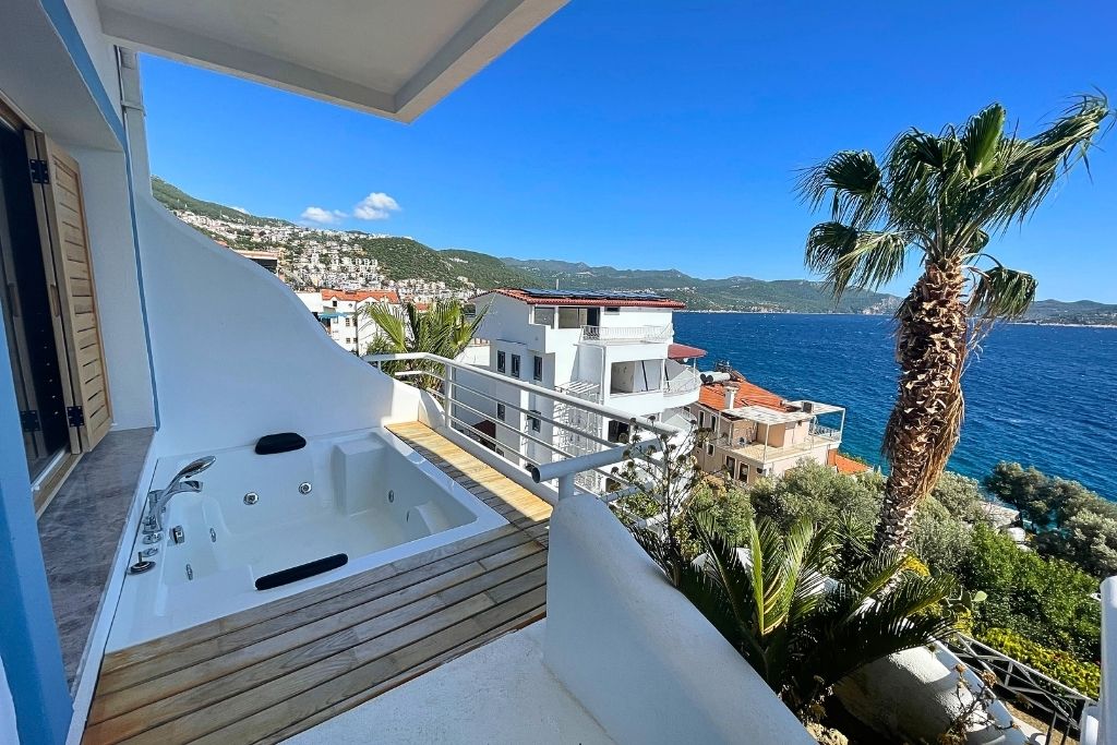 A private balcony hot tub overlooks the deep blue Mediterranean Sea and white hillside buildings in Kaş, Turkey, framed by palm trees and distant mountains.