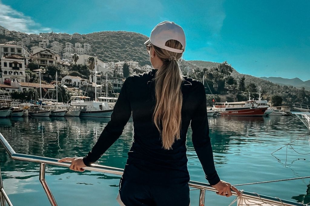 Kate stands at the railing of a boat docked in Kaş harbor, looking out at hillside homes and moored yachts under a bright turquoise sky.