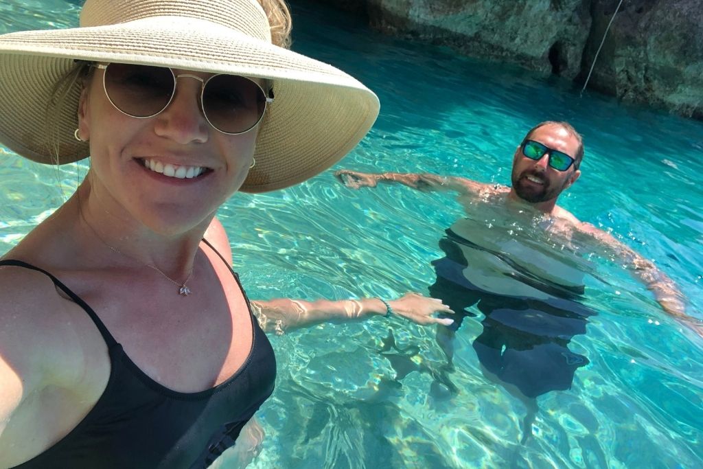 Kate in a sunhat and sunglasses smiles for a selfie while standing in crystal-clear turquoise water, with her husband swimming beside her. They’re enjoying a sunny day in a natural cove, surrounded by rocky cliffs.