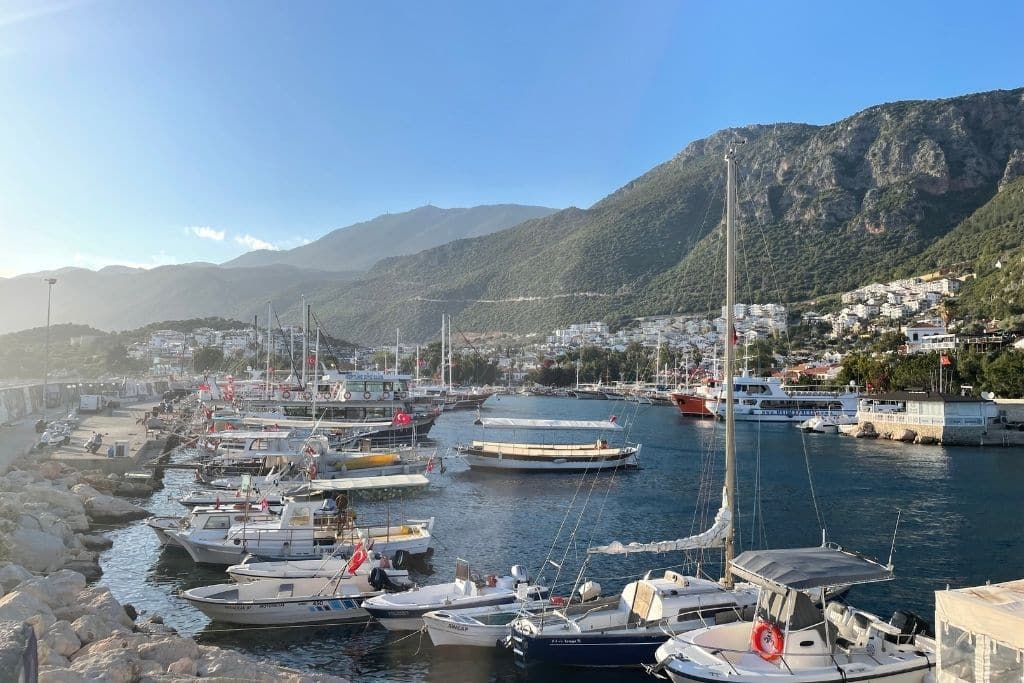 Dozens of boats and yachts line the marina in Kaş, Turkey, backed by lush green hills and white hillside homes glowing in the late afternoon sun.