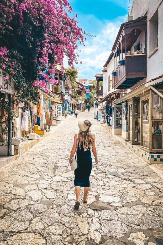 Kate strolls up a cobbled street in Kaş, Turkey, beneath vibrant bougainvillea and flanked by quaint shops and wooden balconies under a bright blue sky.