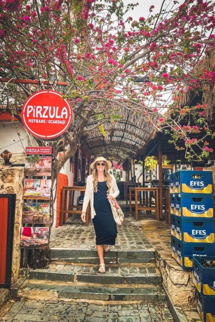 Kate walks past Pirzula restaurant in Kaş, Turkey, under a canopy of blooming bougainvillea with crates of Efes beer stacked nearby.