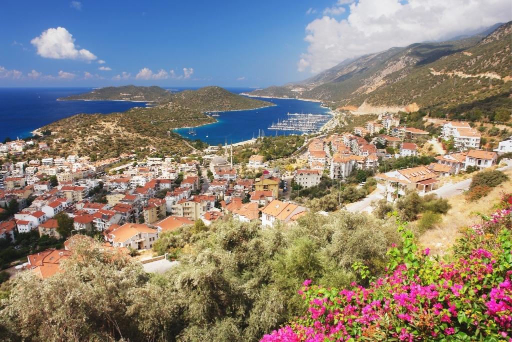Scenic view over Kaş, Turkey, showing red-roofed houses, a marina full of sailboats, and the turquoise coastline framed by green hills and bright pink bougainvillea.