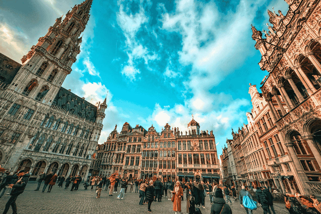 Crowds gather under a bright blue sky at Brussels’ Grand Place, surrounded by stunning Gothic and Baroque buildings, with Kate standing near the center of the square.