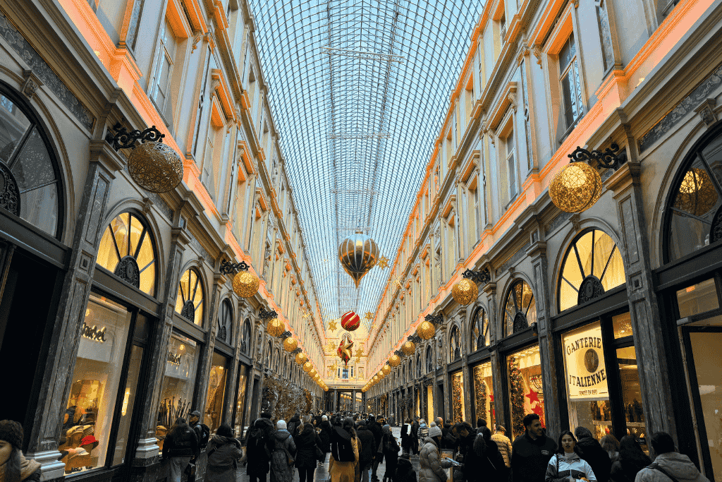 Shoppers stroll beneath golden lights and massive ornaments inside the elegant glass-roofed Galeries Royales Saint-Hubert during the festive season.