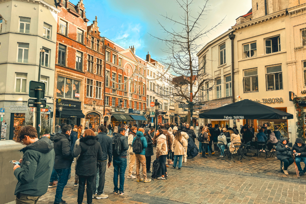 A bustling cobblestone square in Brussels shows crowds gathered near Friterie Tabora, with diners seated under its black canopy.