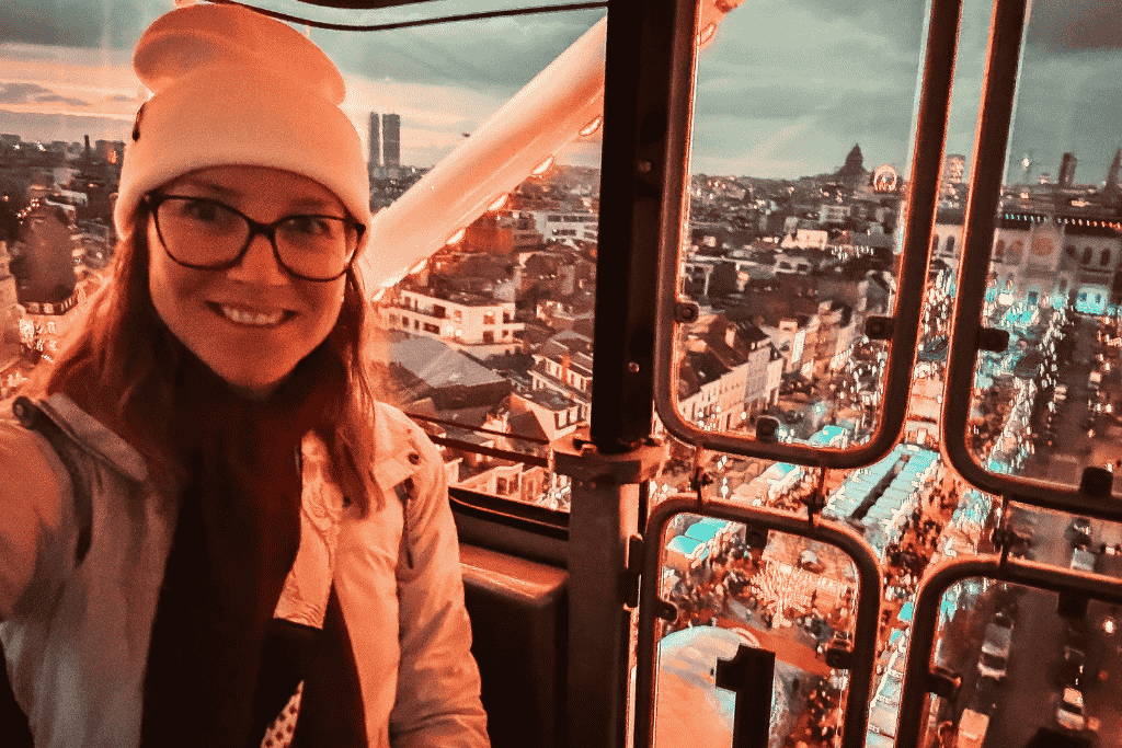 Kate smiles inside a Ferris wheel cabin overlooking the glowing Brussels Christmas market at dusk, with festive lights stretching across the city below.