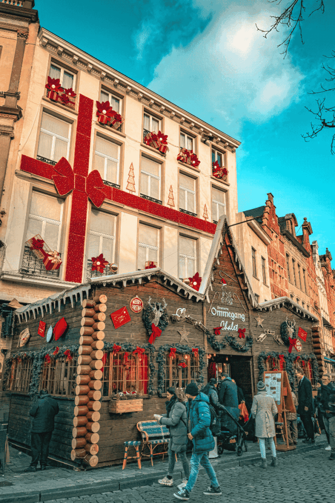 A festive wooden chalet bar named "Ommegang" sits beneath a building wrapped like a giant gift, complete with red bows and holiday decorations in Brussels.