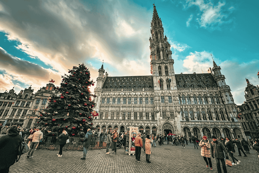 A large Christmas tree decorated with red ornaments stands in Brussels’ Grand Place, with the ornate Gothic Town Hall rising behind it under a dramatic winter sky.
