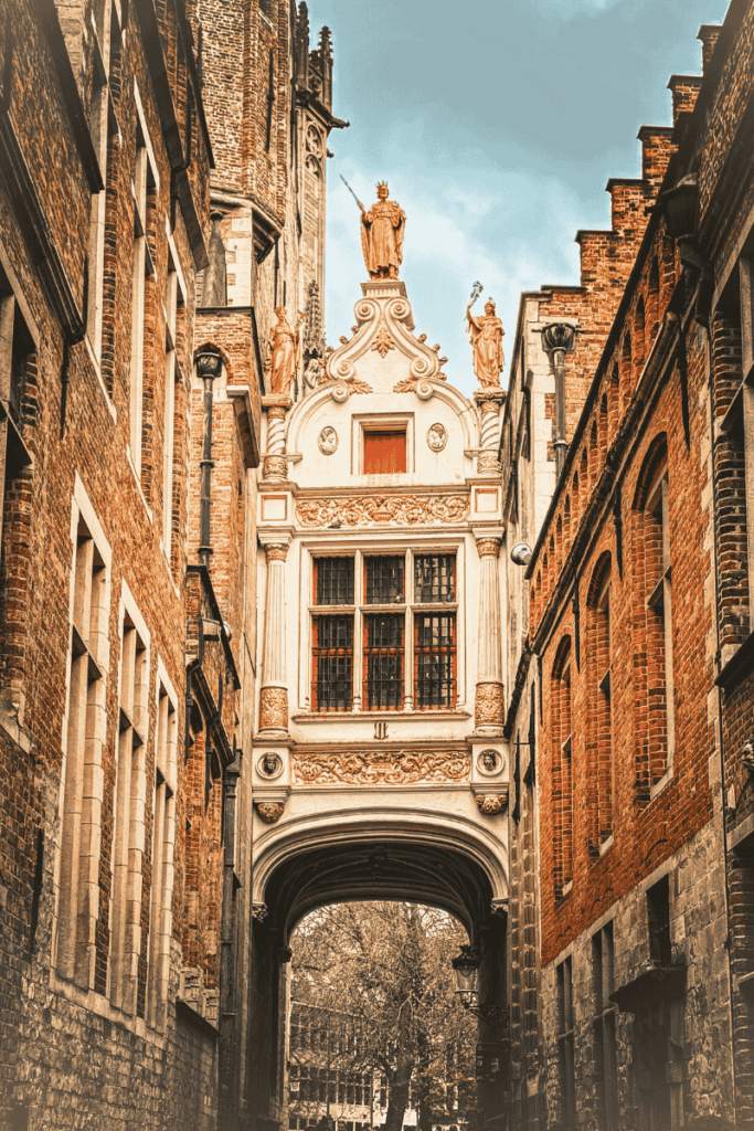 A narrow alley in Bruges frames an ornate white bridge structure topped with gold statues, connecting two historic red-brick buildings.